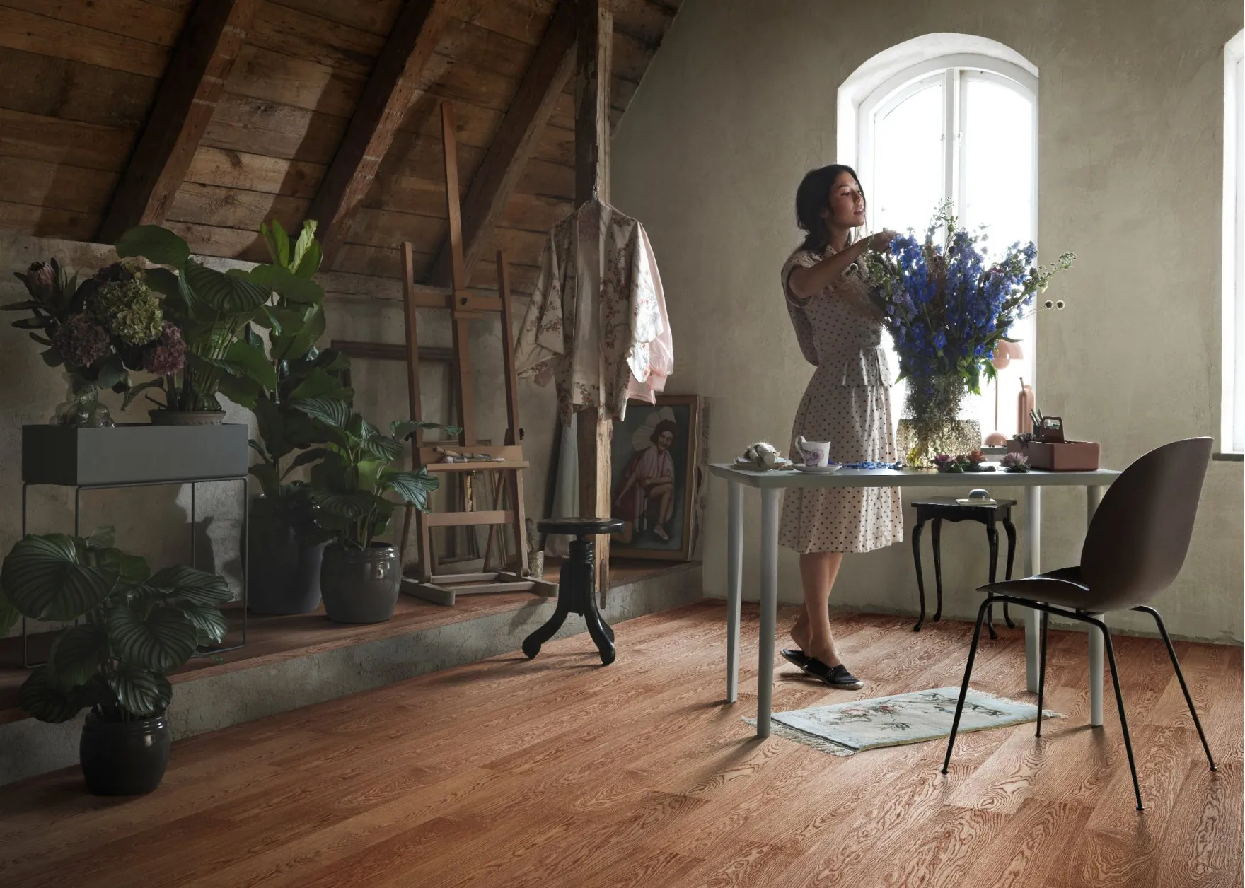 A woman arranging flowers in a sunlit attic room with hardwood floors.