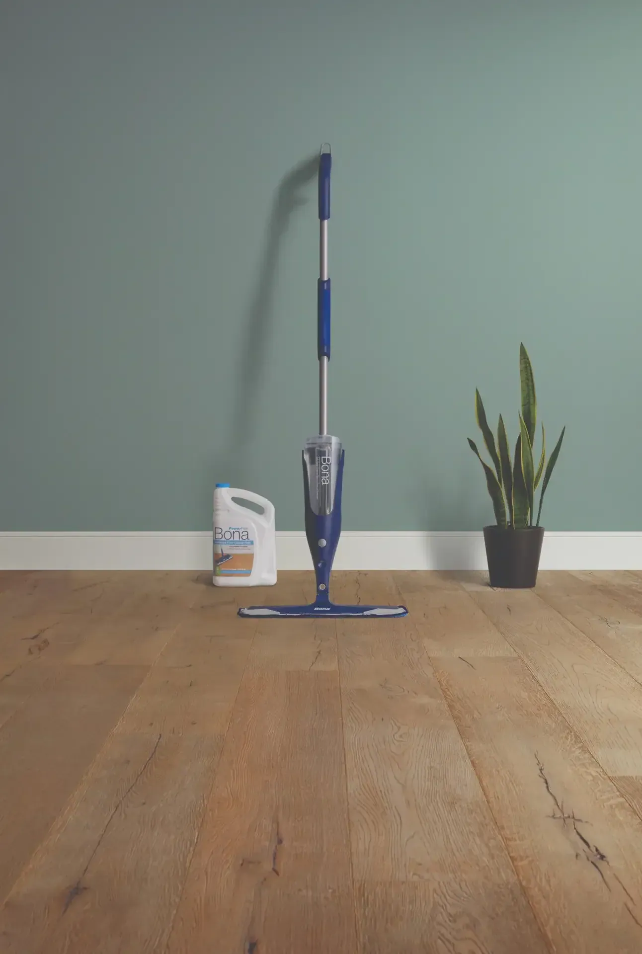 A Bona spray mop leaning against a sage green wall above a hardwood floor.